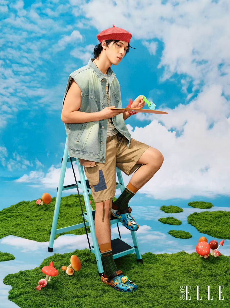 A man wearing a red beret and denim vest sitting on a blue ladder holding a paint palette against a sky and mossy ground backdrop