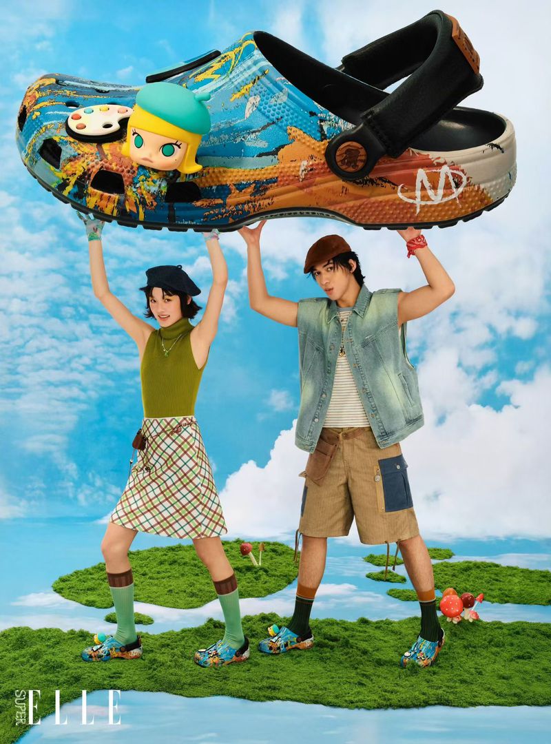 Two models holding up a giant decorated Crocs shoe against a surreal sky and floating mossy islands backdrop.