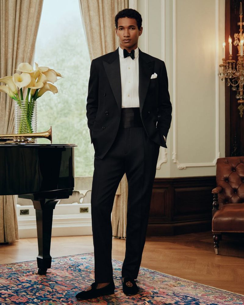 Man wearing a classic black tuxedo standing in a formal room with a piano, floral arrangement, and leather armchair.