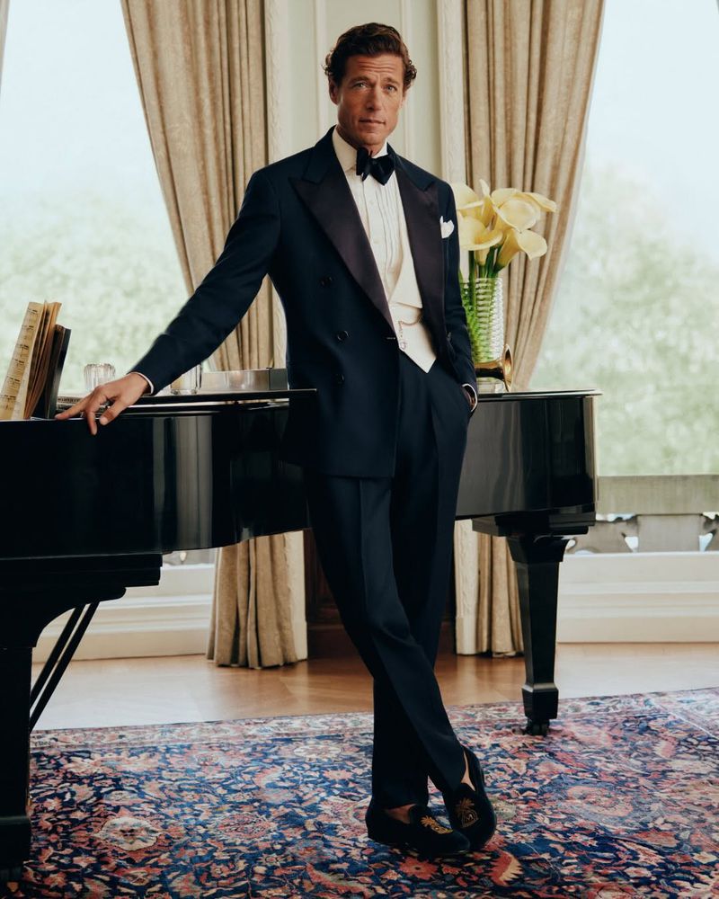 Man wearing a navy tuxedo leaning against a black piano in a classic room