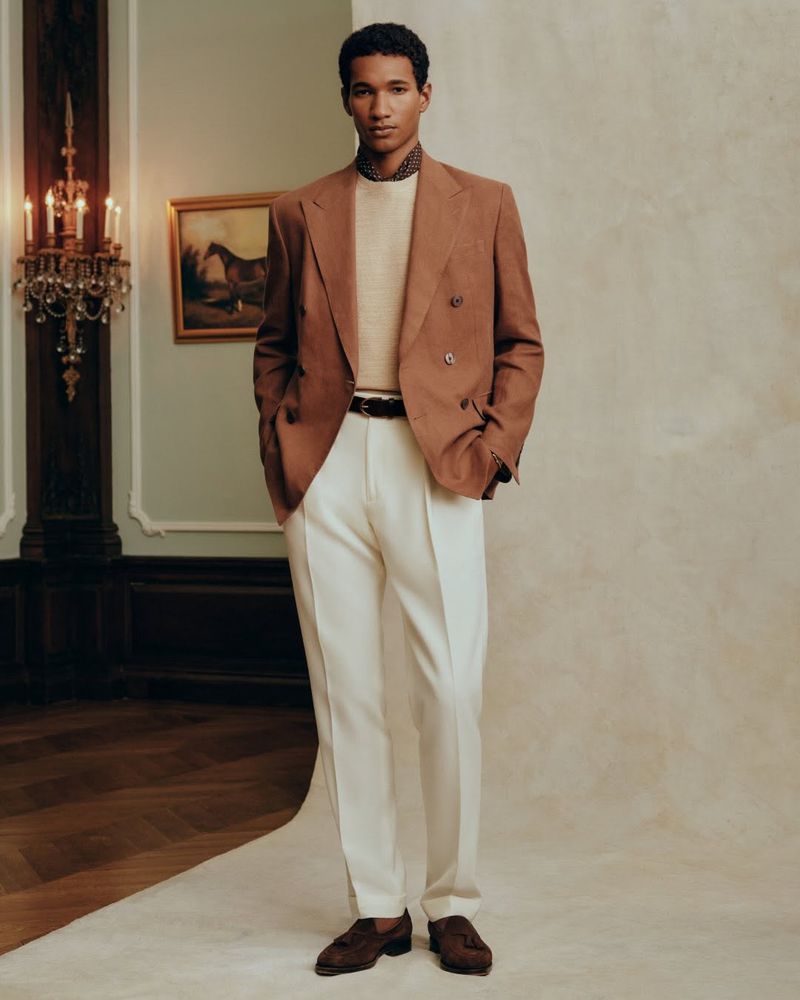 A stylish man in a brown blazer and white pants stands in a classic interior, showcasing a blend of modern fashion and timeless elegance.