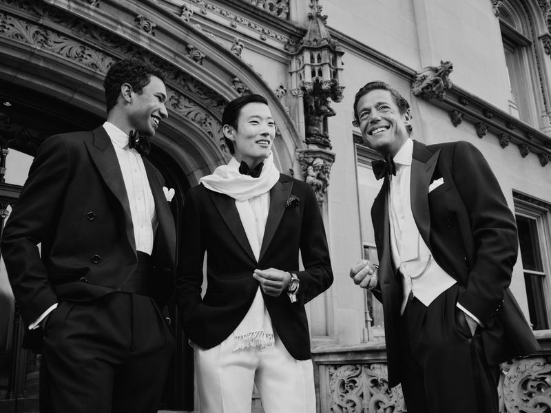 Three male models in formal tuxedos smiling outside an ornate stone building exterior