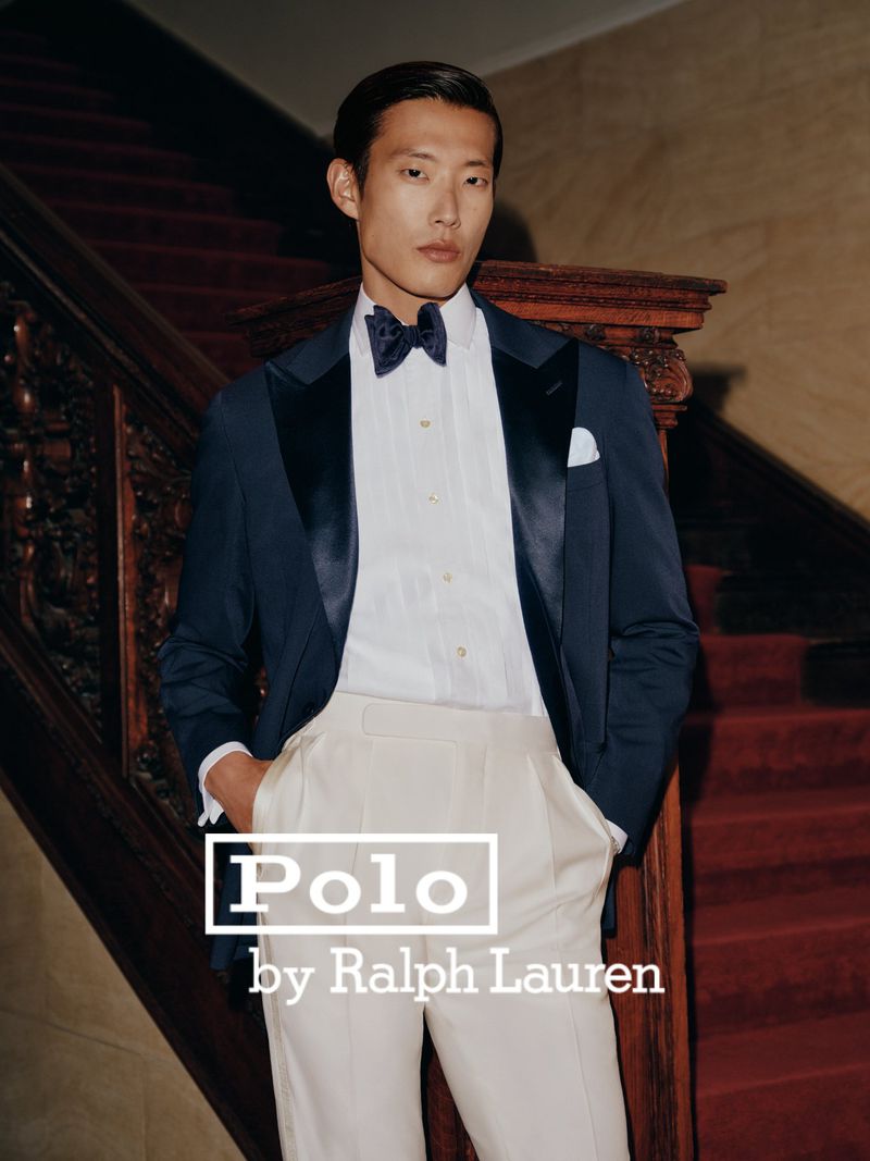 Man wearing a navy tuxedo jacket, white shirt, black bowtie, and cream trousers standing before a grand staircase.
