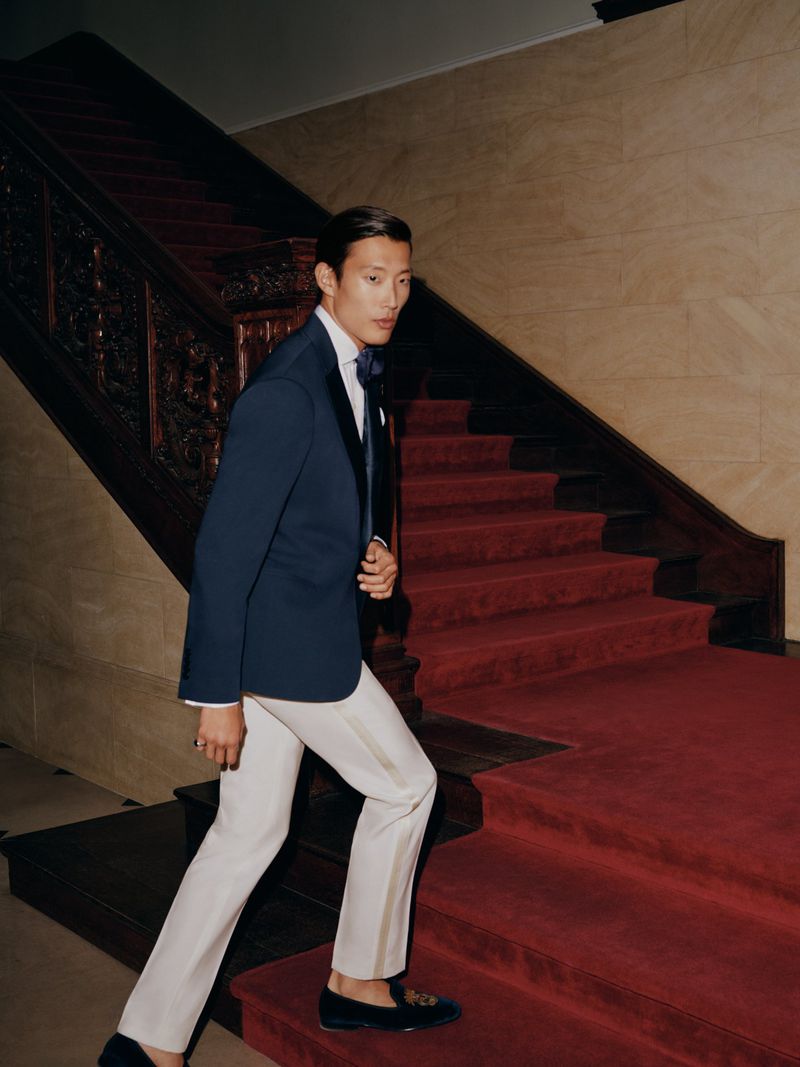 Man wearing a navy tuxedo jacket and white trousers walking up a grand staircase with red carpet