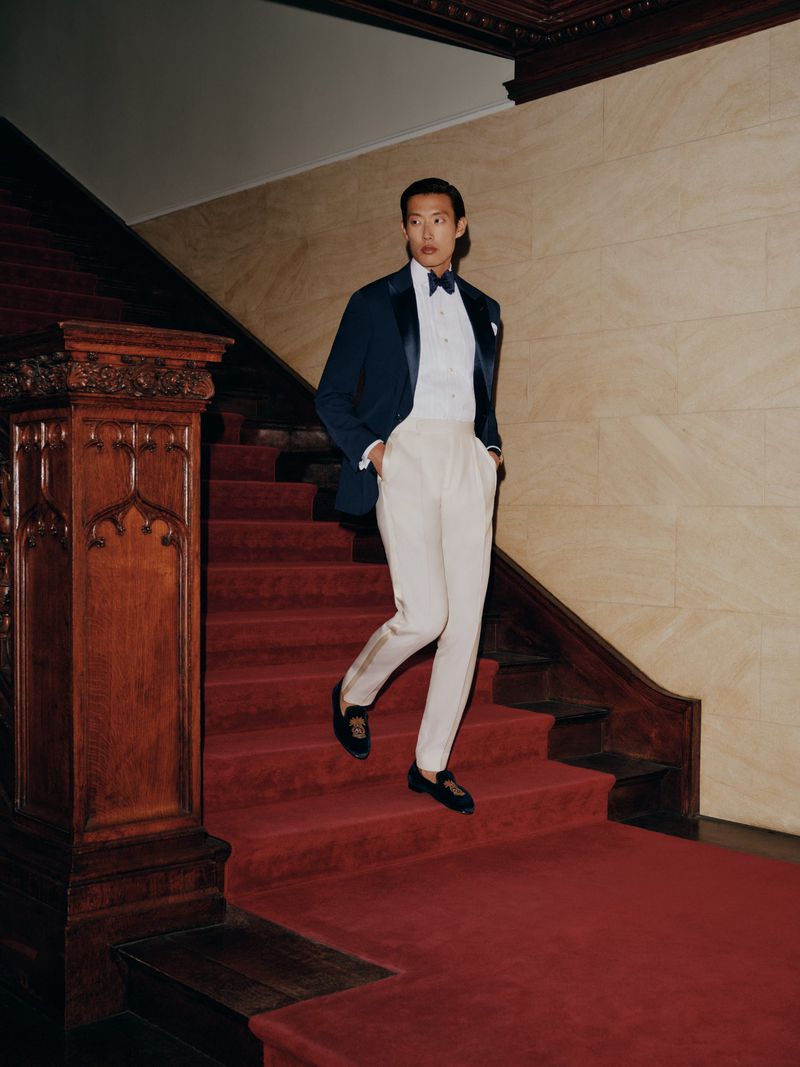 Man wearing a navy tuxedo jacket and cream trousers descending a grand wooden staircase with a red carpet runner.