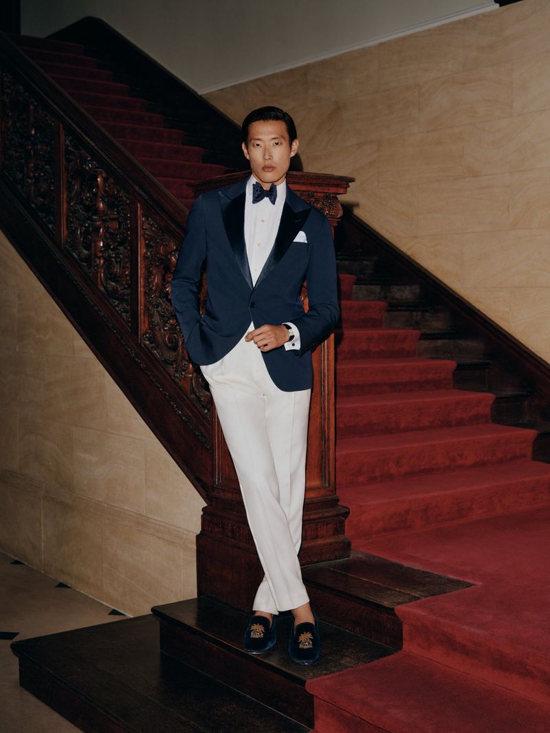 Asian man in navy tuxedo jacket and white trousers standing on a grand staircase with red carpet