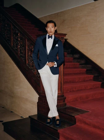 Asian man in navy tuxedo jacket and white trousers standing on a grand staircase with red carpet
