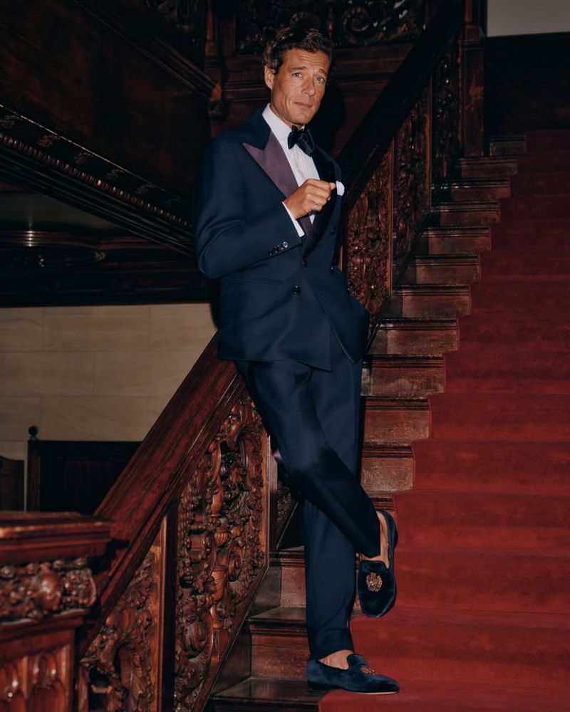 Man in dark suit and bow tie leaning on a grand wooden staircase with a red carpet