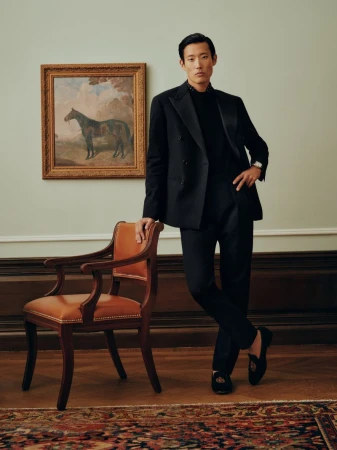 Asian man in a black suit leaning on a wooden armchair against a pale wall with a framed horse painting
