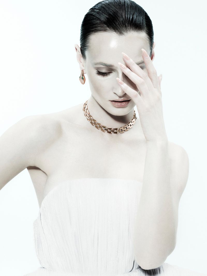 Model wearing a gold link necklace and matching drop earrings against a white studio background, holding a hand near her face.