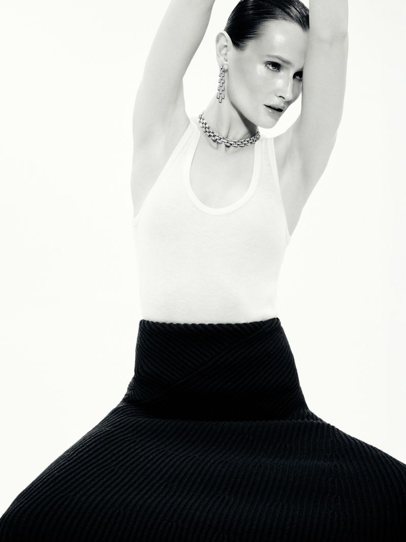 Model in white tank top and black skirt posing against white seamless background with arms raised