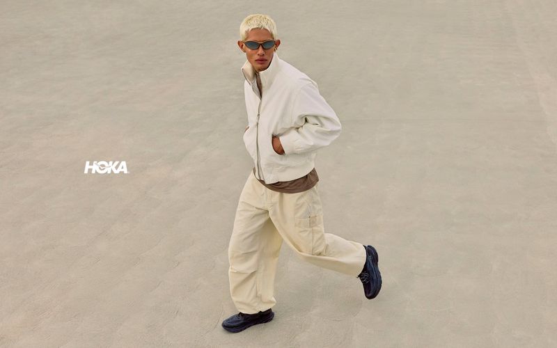 Model with blonde hair wearing white jacket and pants running across concrete background with Hoka logo