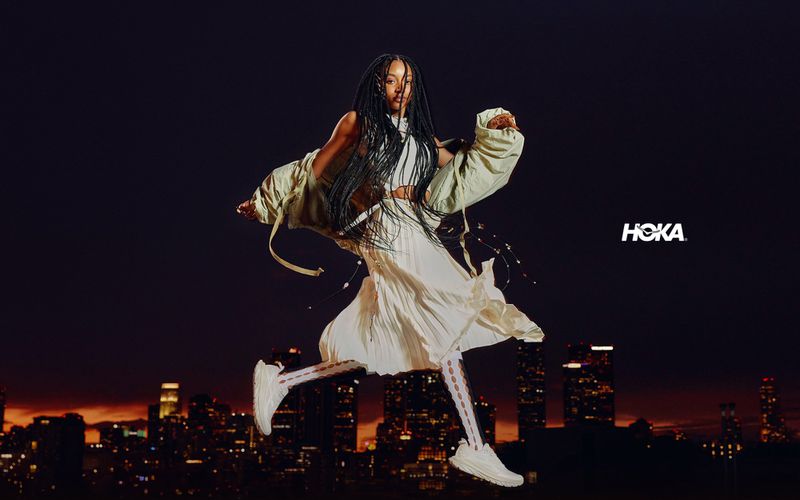 Model in white pleated skirt and sneakers jumping mid-air against a nighttime city skyline backdrop for Hoka Mach Remastered campaign