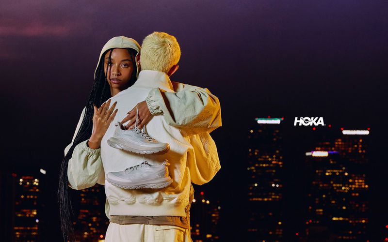 Two individuals in light-colored clothing embrace, displaying two Hoka shoes; a cityscape with the Hoka logo is visible in the background.