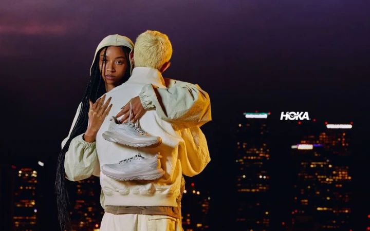 Two individuals in light-colored clothing embrace, displaying two Hoka shoes; a cityscape with the Hoka logo is visible in the background.