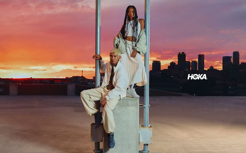 Two models posing on a concrete rooftop at sunset with a city skyline in the background for the Hoka Mach Remastered campaign.