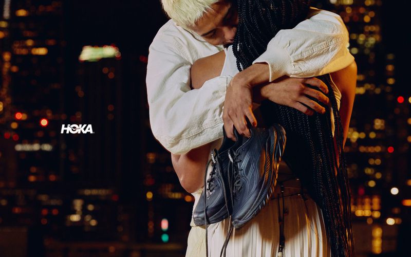 Two people embracing while holding black sneakers against a blurred night city skyline background