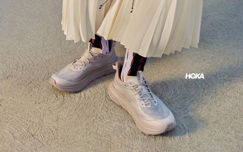 Close-up of pink running sneakers, patterned socks, and a pleated skirt on a textured grey background.