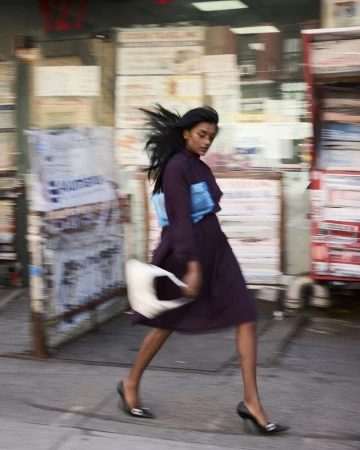 Woman in a purple midi dress walking past a city storefront holding a bright blue pouch and white handbag.
