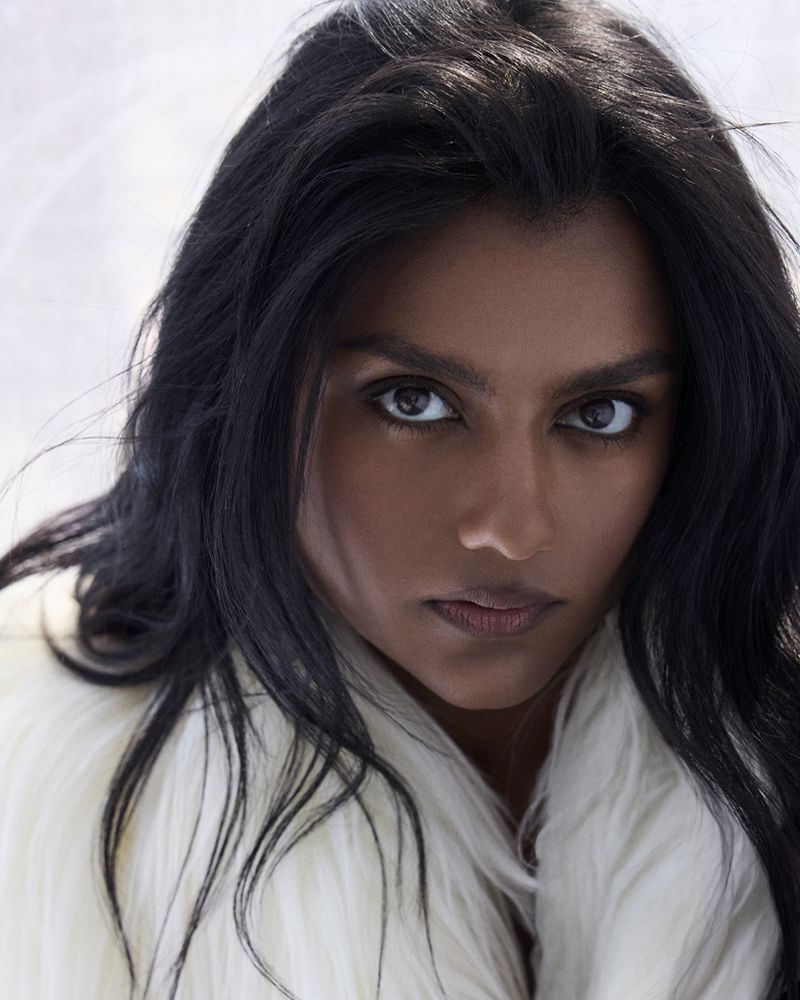 Close-up portrait of a South Asian woman with dark hair wearing a white fur-like jacket, looking directly at the camera with a serious expression.