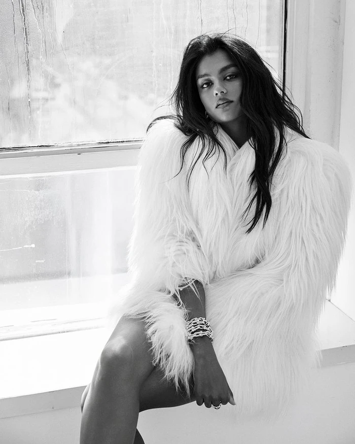 Monochrome fashion portrait of a woman wearing a fluffy white coat seated by a window.