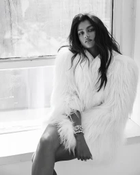 Monochrome fashion portrait of a woman wearing a fluffy white coat seated by a window.