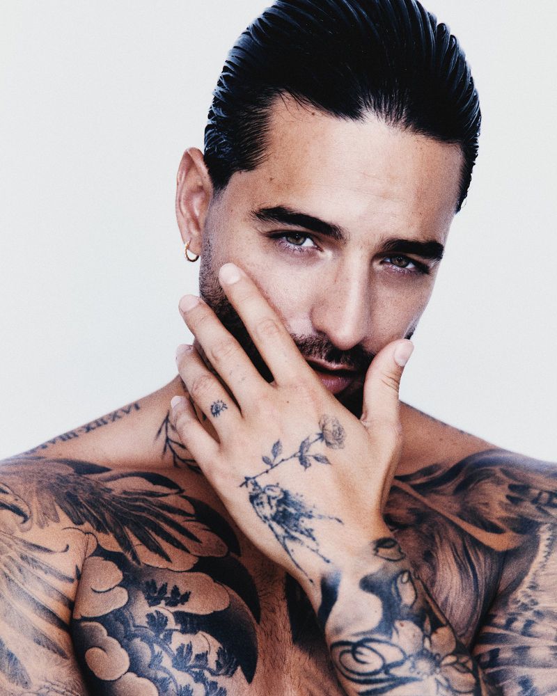 Close-up portrait of a shirtless adult male with tattoos, slicked-back hair, hand near his face, and a neutral expression against white.