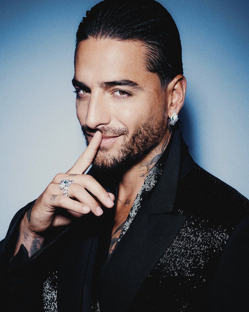 Portrait of Maluma with slicked-back hair, wearing a black jacket and rings, posing against a blue background