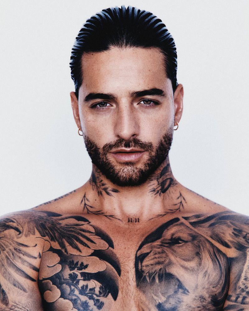 Portrait of Maluma with slicked-back hair and extensive chest and neck tattoos against a solid white background