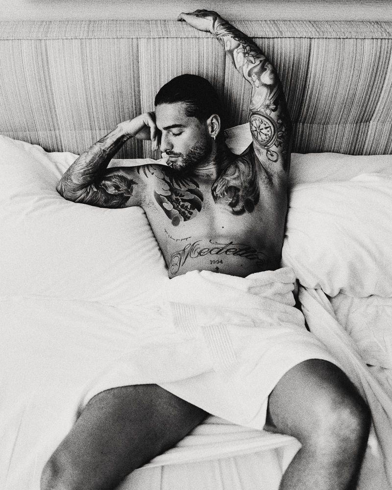 Black and white portrait of singer Maluma lying in bed wrapped in a white towel with tattooed arms and chest.