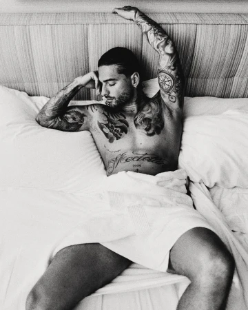 Black and white portrait of singer Maluma lying in bed wrapped in a white towel with tattooed arms and chest.