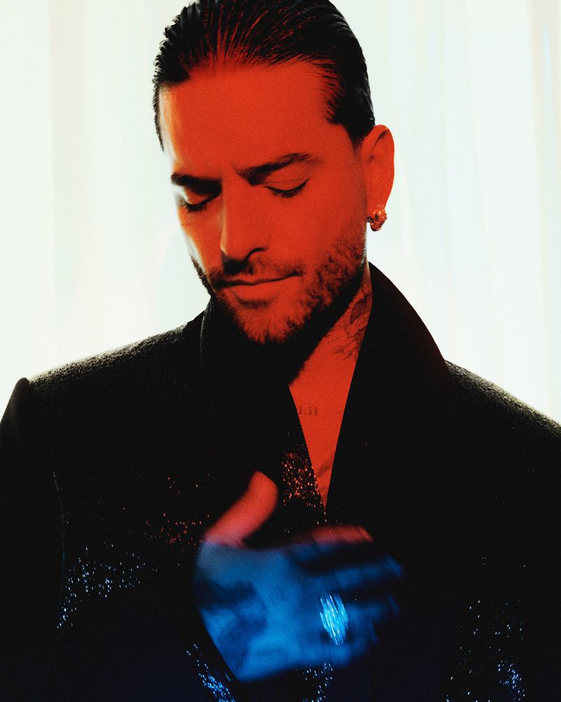 Portrait of Maluma wearing a dark jacket with his hand on his chest under red and blue lighting