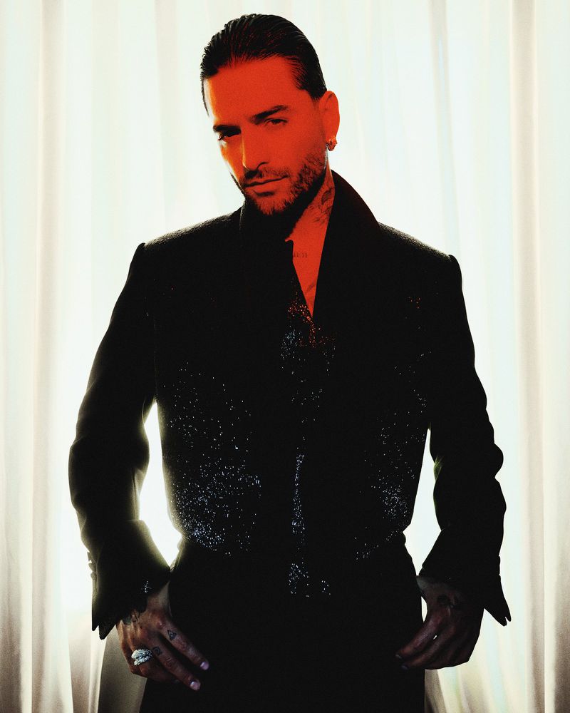 Maluma posing in a dark glittering suit against a sheer white curtain background with warm lighting.