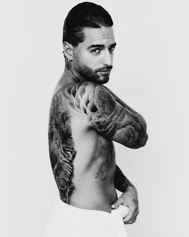Portrait of Maluma with extensive back tattoos and slicked-back hair against a neutral studio background.