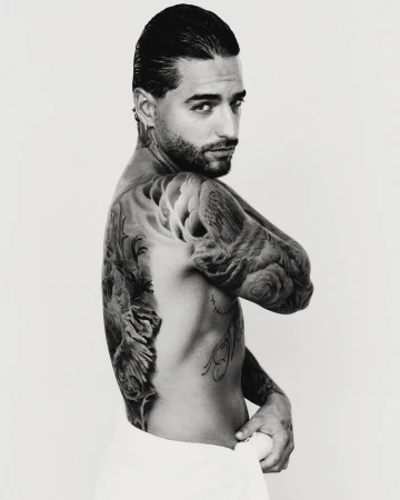Portrait of Maluma with extensive back tattoos and slicked-back hair against a neutral studio background.