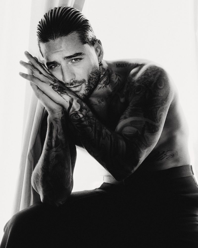 Portrait of Maluma with tattoos and slicked-back hair resting hands against his face against a bright window