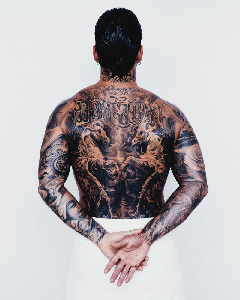 Rear view of a man with extensive upper body and back tattoos wrapped in a white towel against a seamless background.