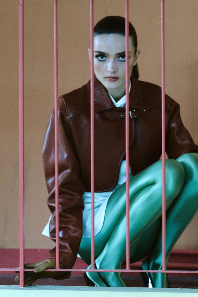 A fashion shot of a woman in a brown jacket and green tights crouching behind pink bars, looking directly at the camera with striking makeup.