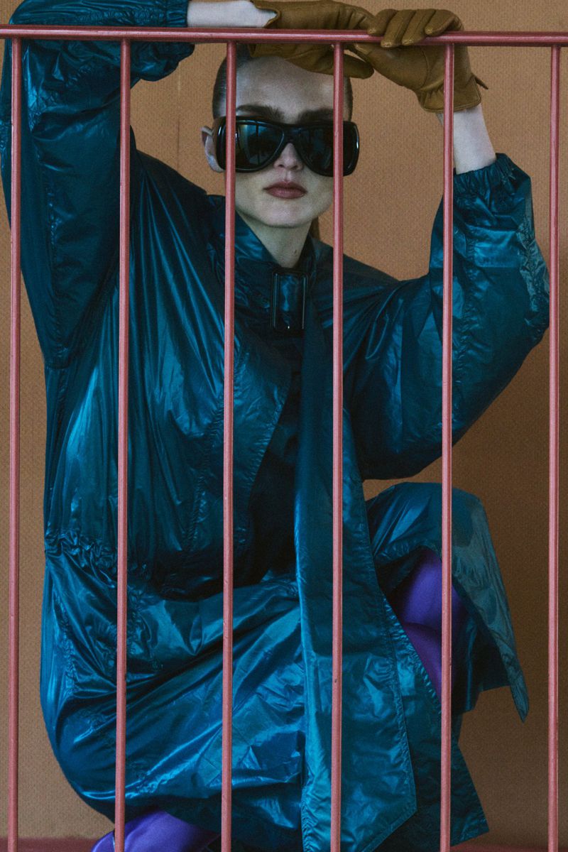 Model Gertrud Rose in a blue windbreaker wearing dark sunglasses and brown leather gloves behind pink vertical bars.
