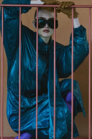 Model Gertrud Rose in a blue windbreaker wearing dark sunglasses and brown leather gloves behind pink vertical bars.