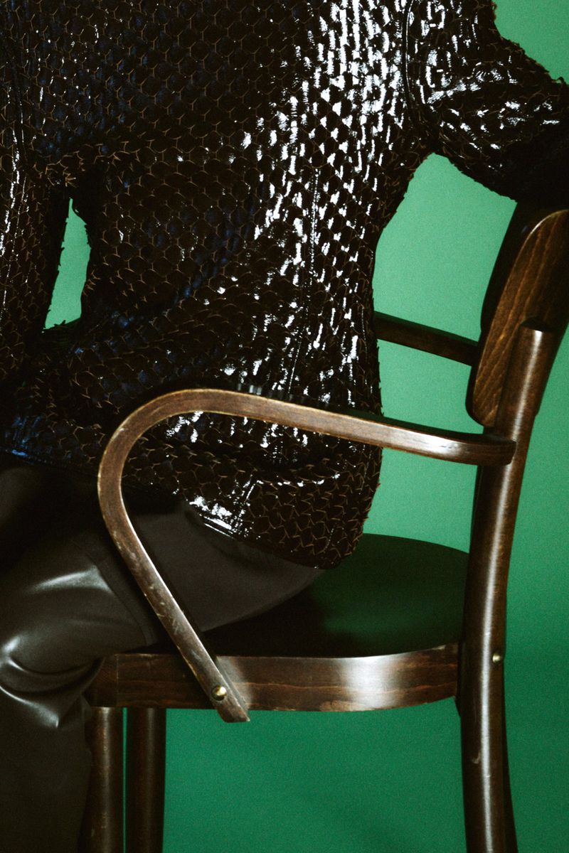 Model Gertrud Rose seated on a wooden chair wearing a textured dark jacket against a bright green background
