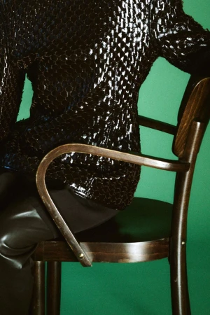 Model Gertrud Rose seated on a wooden chair wearing a textured dark jacket against a bright green background