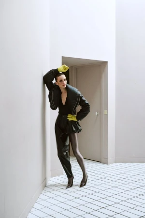 Model Gertrud Rose leaning against a wall wearing a dark puffy jacket and neon green gloves in an indoor architectural corridor.