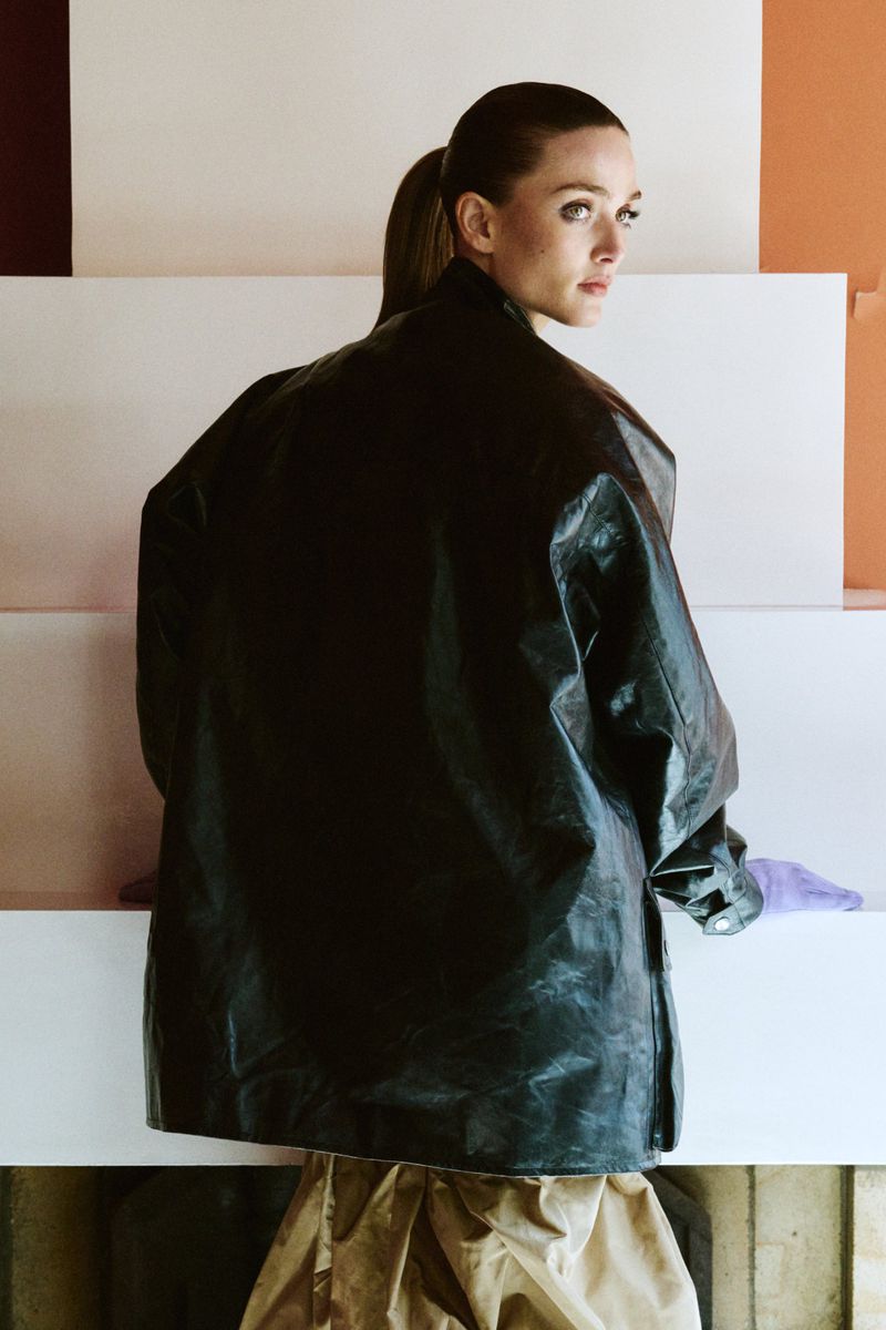Model Gertrud Rose wearing an oversized dark coat and standing against a structured architectural background.