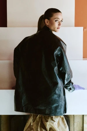 Model Gertrud Rose wearing an oversized dark coat and standing against a structured architectural background.