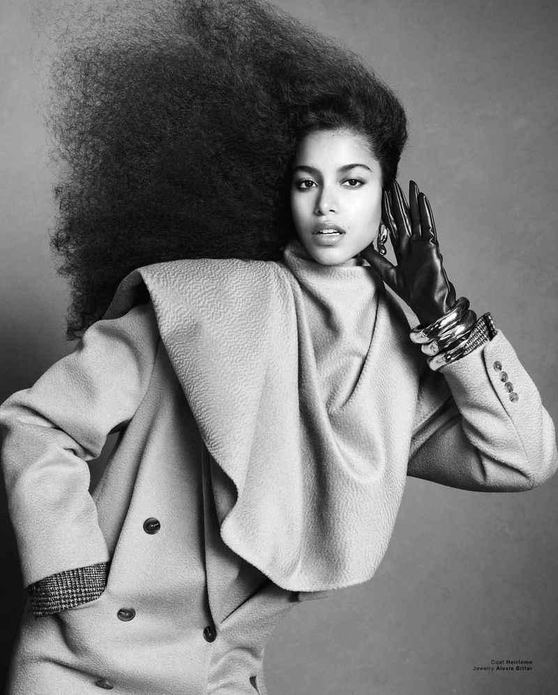 Model Luna Yohannan with voluminous dark hair wearing a textured beige coat and black leather gloves in a black and white editorial portrait.