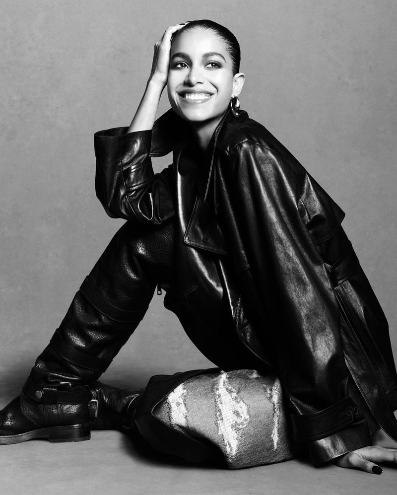 Model Luna Yohannan seated wearing a black leather coat and tall boots against a neutral grey studio background