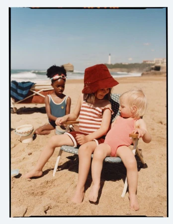 Three young children sitting on small beach chairs on a sandy beach with the ocean and a distant lighthouse in the background.