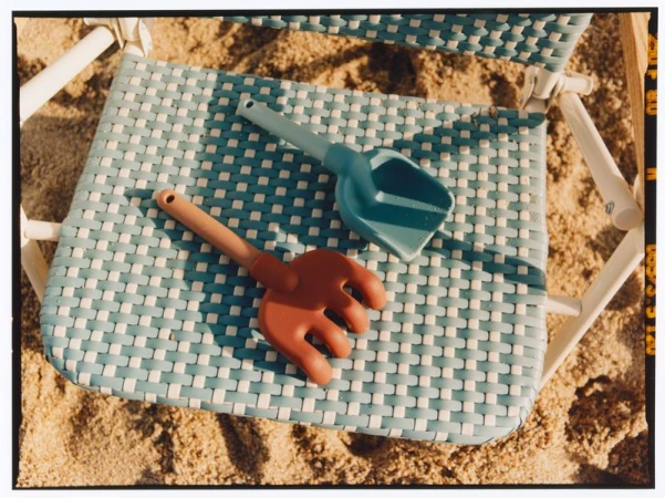 Blue plastic shovel and brown rake resting on a woven beach chair over sand.
