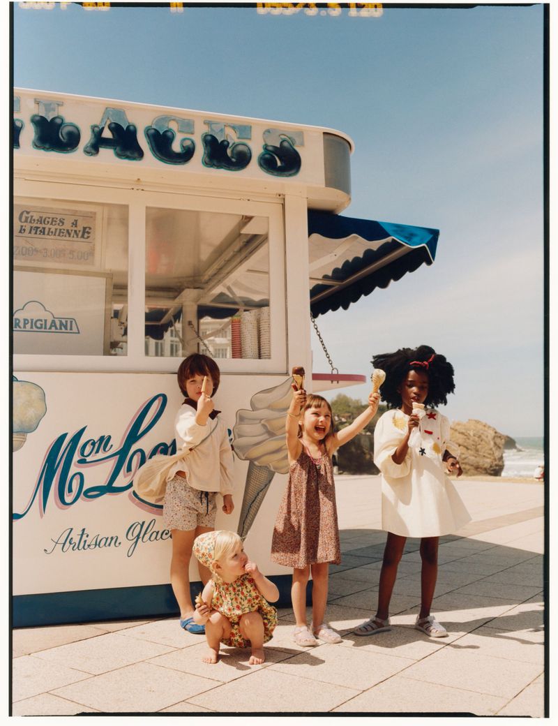 Four children holding ice cream cones in front of an ice cream kiosk near the ocean under a clear sky.